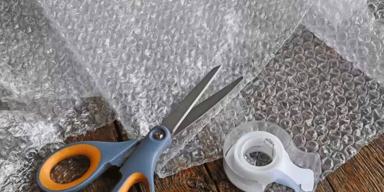 Packaging prep: bubble wrap with air filled bubbles, scissors, and tape on a rustic wooden table.