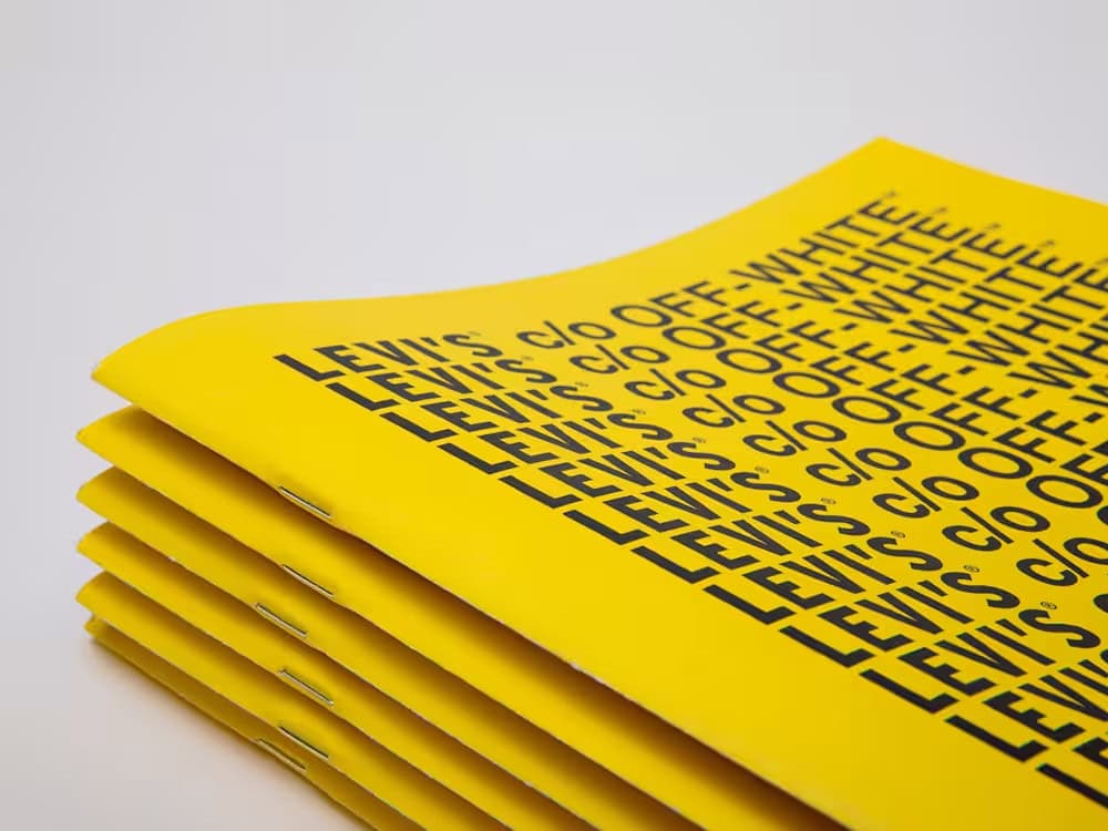 Pile de livrets jaune vif agrafés, portant l'inscription répétée « LEVI'S c/o OFF-WHITE », avec des agrafes métalliques visibles sur le bord gauche, présentés sur un fond blanc uni.