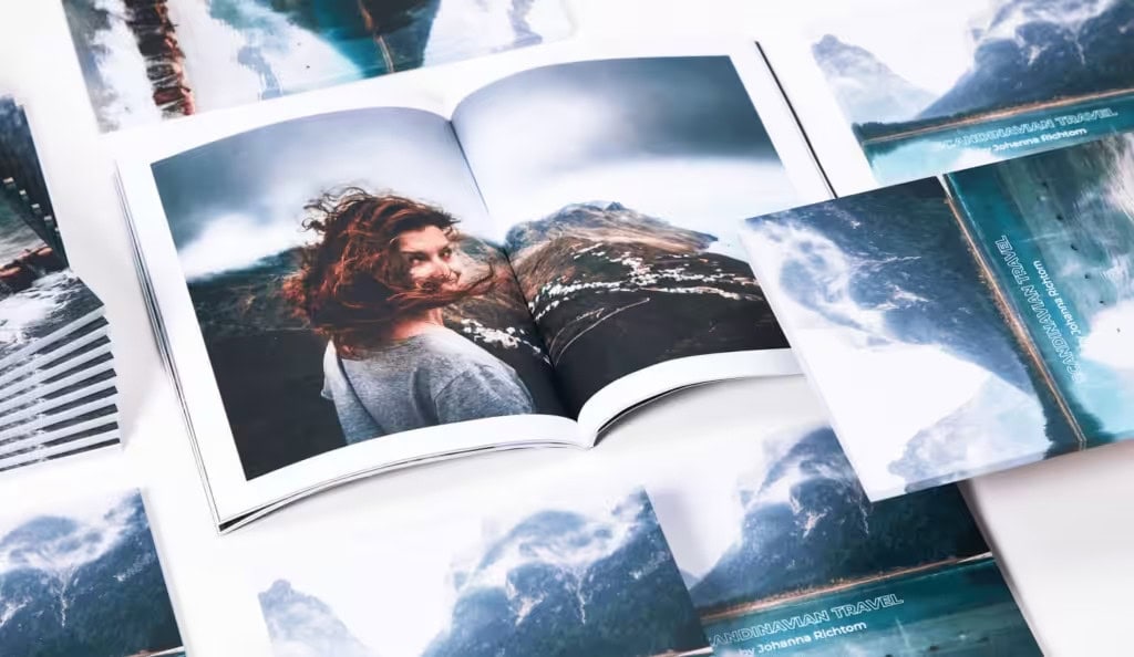 Open perfect-bound travel photobook titled "SCANDINAVIAN TRAVEL" lies flat, displaying landscape and portrait photos, with additional matching booklets scattered around on a white surface.