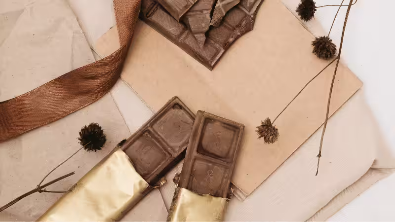 Flach arrangiert sind Schokoladenriegel in umweltfreundlicher Kraftpapier- und Goldfolienverpackung, dekoriert mit Trockenblumen und einem braunen Band.