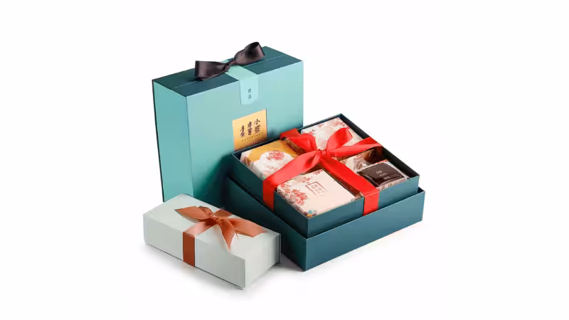 Elegant teal gift box with red ribbon, open to reveal branded packages. Includes a smaller white box with brown bow. Minimalist product shot on white background.