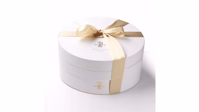 Luxury round white gift box with gold satin ribbon bow, embossed with gold brand logo, minimalist product shot on white background.