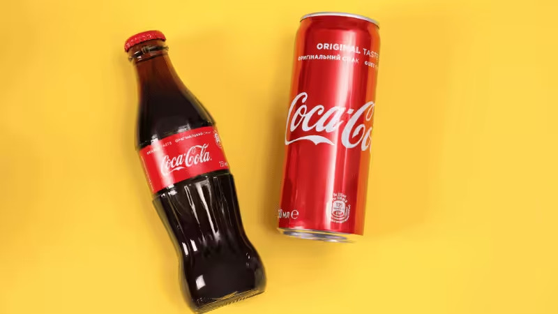 A glass bottle and a can of Coca-Cola with red labels are placed on a bright yellow background. The bottle is lying slightly tilted, and the can is standing upright.