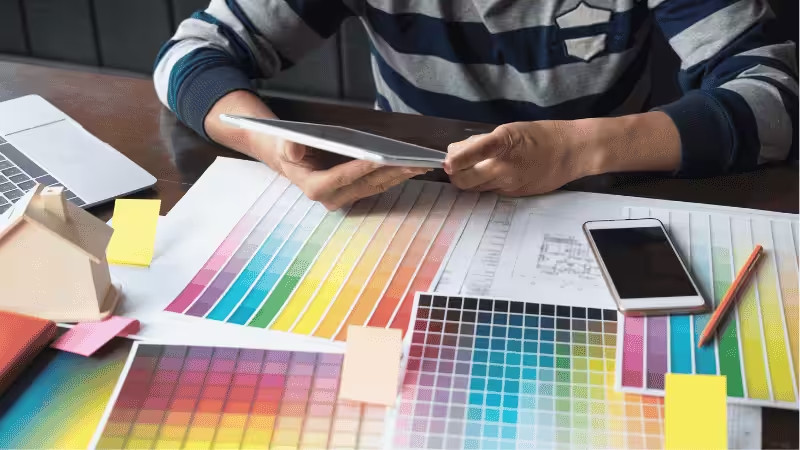 Person using a tablet at a desk with color swatches, a smartphone, pencil, sticky notes, and a laptop, suggesting design or color selection work.