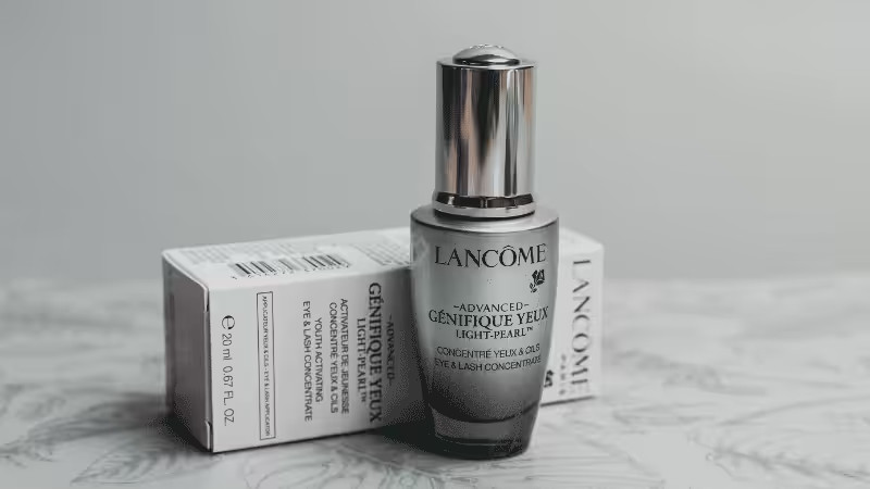 A bottle of Lancôme Advanced Génifique Yeux Light-Pearl eye serum stands in front of its packaging box on a light patterned surface.