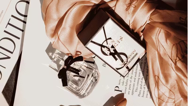 A YSL perfume bottle and box lie on open magazines, accented with a sheer peach scarf, forming an elegant, luxurious flat lay.