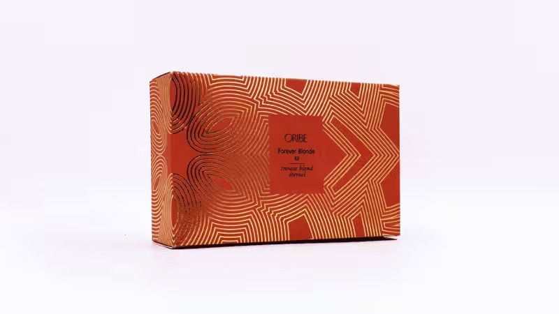 Rectangular orange box with gold geometric swirls and lines. Center label reads “ORIBE Forever Blonde & treasure found eternal” on a plain white background.