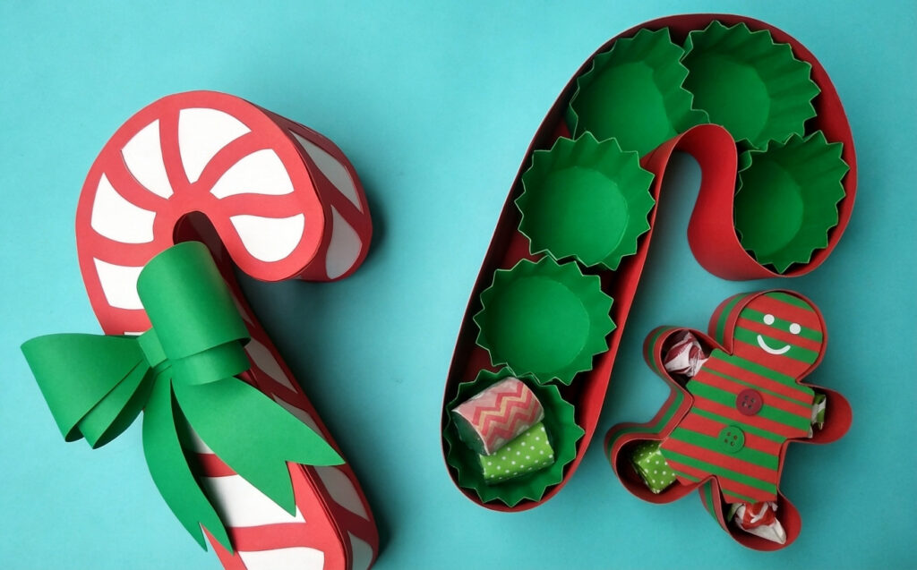 Open red candy cane-shaped gift box with green liners, plus a gingerbread man treat box, on a turquoise background.