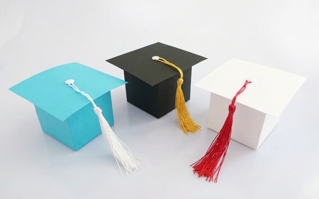 Three graduation cap-shaped gift boxes in turquoise, black, and white, each with a matching tassel, on a white background.