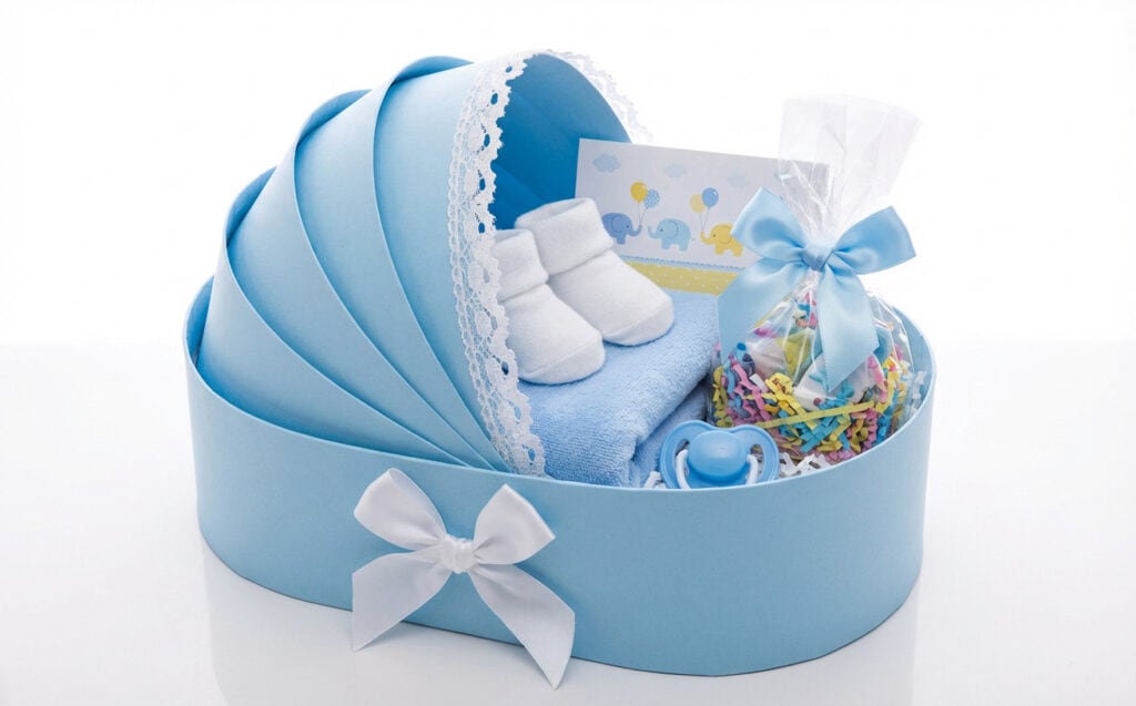 Charming blue baby bassinet-shaped gift box adorned with white lace and satin bows, filled with baby essentials including a pair of white booties, a blue pacifier, and colorful gift grass.