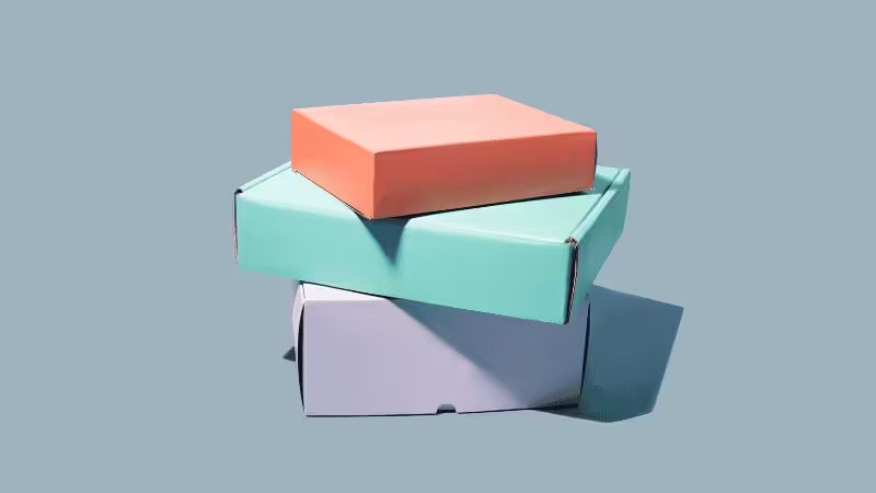 Three cardboard boxes stack: coral orange top, teal middle, light gray bottom. Plain background, shadows cast to their right, simple clean product-style presentation.
