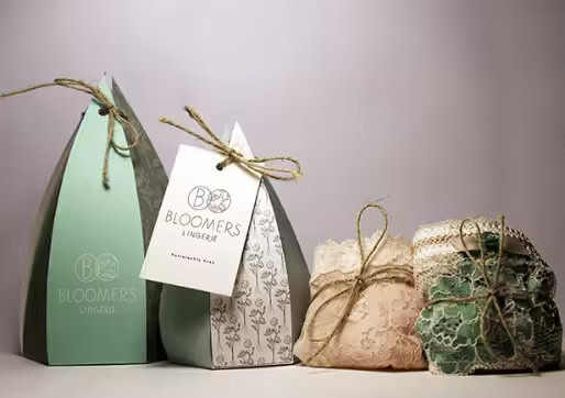 Assortment of uniquely shaped Bloomers Lingerie gift boxes: pyramid paper boxes with twine, and lace-wrapped pouches, on a neutral background.