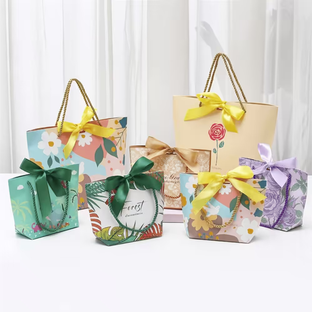 Assorted decorative gift bags and gift boxes in vibrant floral, tropical, and rose prints, tied with satin bows and rope handles.