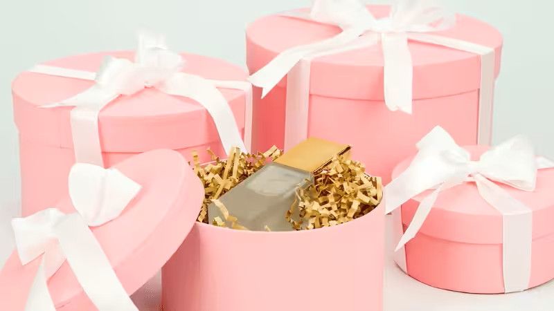 Elegant pink round gift boxes for women, with one open revealing a perfume bottle in gold crinkle paper filler.