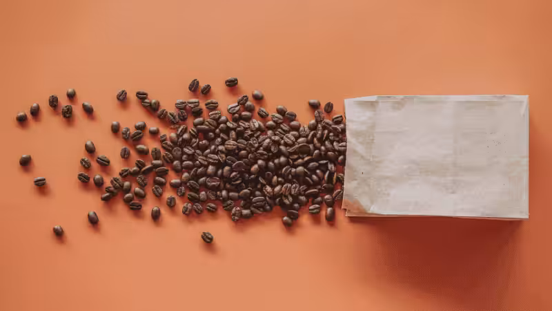 Roasted coffee beans spilling from an open white kraft paper bag, flat lay on a warm terracotta orange background.