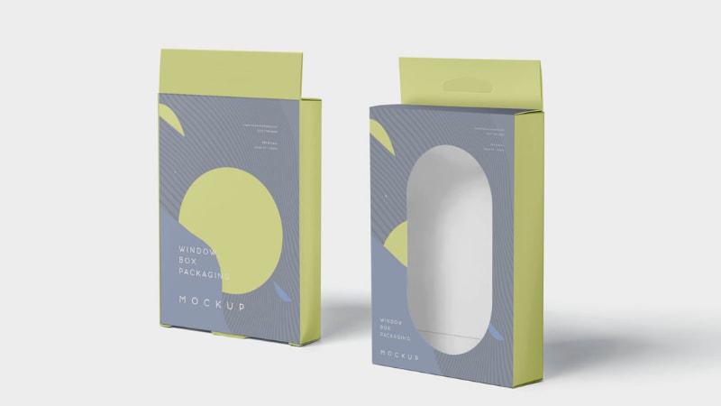 Two 3D window box packaging mockups with grey-green geometric designs, one closed and one with an oval window, on a light gray background.