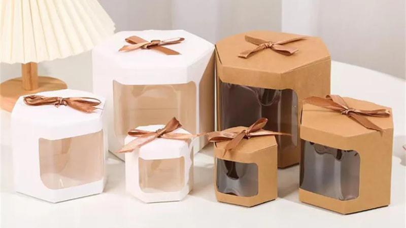 Set of hexagonal window box packaging in white and kraft brown, adorned with brown ribbons, featuring clear windows to showcase contents.